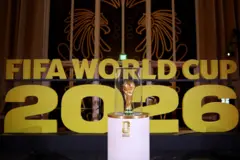 Le trophée de la FIFA entouré de verre transparent, posé sur une table ronde, avec au fond un écrit : "Fifa World Cup 2026" (la Coupe du Monde de la FIFA 2026).