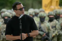 El presidente de Ecuador, Daniel Noboa, frente a un micrófono y frente a un grupo de soldados. Viste una franela negra y lleva gafas de sol negras.