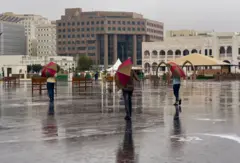 يتجول 3 أشخاص، بمظلات في سوق واقف، خلال يوم ماطر في الدوحة- قطر. تظهر مجموعة من المباني، والأمطار تشكل تجمعاً من المياه.