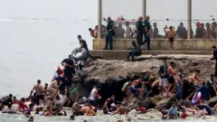 Egy csoport ember kapaszkodik fel a tengerből a sziklákra. Többen még a vízben vannak, úszóeszközök segítségével. A sziklák tetején drótkerítés húzódik.