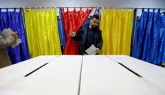 Мужчина выходит из кабинки для голосования с занавесками цветов румынского флага - темно-синими, желтыми и красными - и несет бюллетень к избирательной урне.