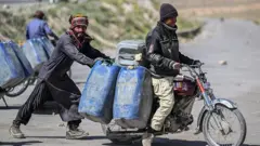 Um homem empurra uma motocicleta enquanto outro está sentado. A motocicleta transporta grandes galões de combustível