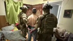 Двое мужчин в камуфляже, бронежилетах и касках держат под руки обнаженного по пояс мужчину, у которого руки за спиной скованы наручниками. На стене в комнате виден ковер и зеленая занавеска