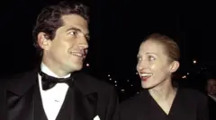 John F Kennedy Jr, vistiendo un traje de etiqueta, junto a Carolyn Bessette-Kennedy, con un vestido negro de cuello en v. Ambos sonríen, mientras parecen conversar.