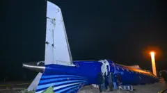 Несколько мужчин осматривают хвост самолета, потерпевшего крушение. Хвост перевернут сверху вниз, на нем видны повреждения неясной природы.