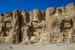 Antiga necrópole persa escavada diretamente em uma enorme parede rochosa. A pedra calcária apresenta portais com formas complexas. Turistas passam caminhando sob o céu azul intenso.