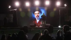 Foto do novo líder supremo do Irã, Mojtaba Khamenei, iluminada no escuro em uma tela em Teerã e exibida para a multidão. Um homem está de pé em frente à fotografia, com um microfone e um alto-falante, aparentemente se dirigindo à multidão