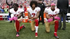 Colin Kaepernick (hagati) n'abandi bakinyi bagenziwe babiri bapfukamye igihe ururirimbo rw'igihugu rwa Amerika rwariko rusamirana mu 2016