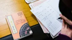 Uma mão com um lápis escrevendo em um caderno com anotações. Ao lado há um livro de astrologia