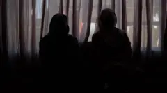 Duas mulheres se sentam no escuro usando hijabs e máscaras faciais para proteger sua identidade