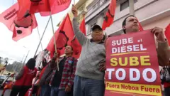 Manifestantes gritam com cartazes dizendo: "Sube el diésel, sube todo. Fuera Noboa Fuera" numa rua de Quito