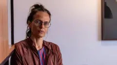 Foto de uma mulher de óculos de grau, cabelos presos, vestindo uma jaqueta e olhando para a câmera.