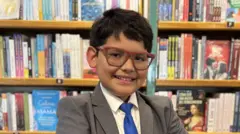 David Camacho, menino com sorriso largo, óculos, com terno cinza e gravata azul, posa em frente à estante de uma livraria