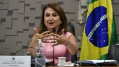 Kátia Abreu gesticula, sentada diante de mesa de comissão, com bandeira do Brasil atrás
