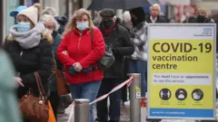 Pessoas agasalhadas e com máscaras em fila em área externa, ao lado de placa dizendo: Covid-19 Vaccination Centre