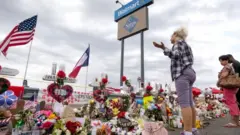Três pessoas em frente a flores e outras demonstrações de homeangem em área externa do local onde ocorreu ataque em El Paso