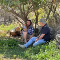 Jeremy Bowen (R) e Meir Simcha sentados na grama sob uma figueira