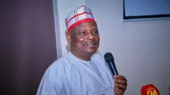 Leader of di NNPP Rabiu Kwankwaso wear white agbada, red and white cap and e hold microphone 