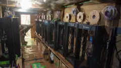 Impressoras 3D enfileiradas sobre uma mesa, com um homem vestido com uniforme militar supervisionando uma delas