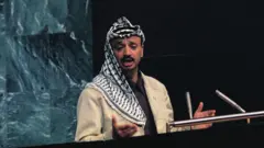 Yasser Arafat habla desde el podio de oradores. Viste un chaqueta beige, una camisa negra y lleva sobre la cabeza una kufiya palestina.