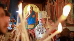 Celebração sendo realizada na igreja por um padre. Pessoas seguram velas