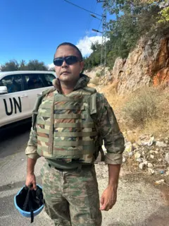 Hamilton de Andrade dos Santos de uniforme, em pé, segurando capacete em uma estrada vazia, ao lado de um carro com os dizeres 'UN'