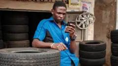 Umugabo wambaye ishati y'ubururu, uhugiye muri telefoni igendanwa