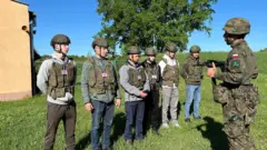 Seis pessoas usando capacetes e coletes camuflados assistem a um soldado, também em uniforme de camuflagem com a bandeira da Polônia, demonstrar uma arma. Estão posicionados em um campo aberto
