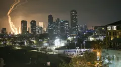 以色列特拉維夫的夜空中，有導彈劃過天空的煙霧，遠處地面的爆炸造成的火光。