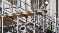 Građevinska skela i dva radnika u prslucima i sa kacigama na glavi