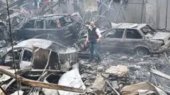 мужчина стоит посреди руин дома и остовов сгоревших машин в Бейруте