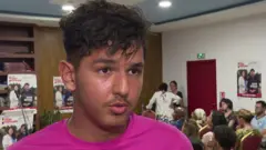 Mehdi Kessaci hablando con alguien fuera de cámara con pósteres de su hermano al fondo