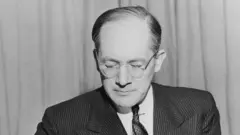 Uma foto em preto e branco mostra o advogado judeu polonês Raphael Lemkin, vestido com um terno completo, sentado à mesa com um microfone, caneta na mão, enquanto escreve em uma folha de papel.