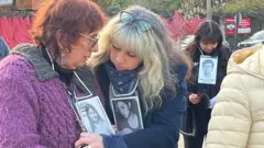 En la imagen, se ve a Holzapfel, a la izquierda, junto a otra mujer rubia a su derecha. Las mujeres se encuentran frente al monumento conmemorativo y llevan colgadas al cuello fotografías en blanco y negro de personas desaparecidas y antiguas víctimas del régimen de Pinochet.