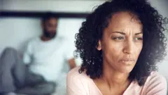 Mulher com semblante triste. Seu marido aparece ao fundo, desfocado