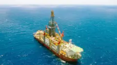 Navio-plataforma de perfuração offshore visto de cima, cercado pelo mar, com uma grande torre central, guindastes e heliponto na popa.