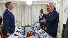 Uma fila de homens de terno em pé ao redor de uma longa mesa com garrafas de água de plástico e microfones finos, em frente a um homem que está sozinho à mesa