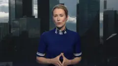 Umugore w'umu blonde mu mpuzu y'ubururu inigirije mw'izosi ariko aravuga araba camera micro bayifatishije kw'ikoro. Ariko aravuga yageranije ibiganza