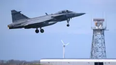 JAS-39 Gripen на взлете: нос самолета направлен по диагонали вверх, за ним видны белые лопасти ветряной электростанции и решетчатая башня аэродрома с антеннами