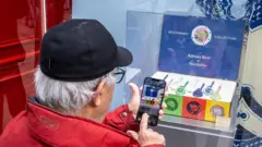 Um homem grisalho com uma jaqueta vermelha e boné preto tira uma foto com seu smartphone de uma vitrine de relógio Royal Pop Swatch em Paris.
