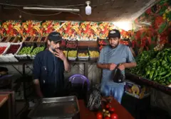 Dois homens estão parados no centro de um supermercado. Um deles, à esquerda, vestido de azul escuro, usa boné e fuma um charuto. O outro, à direita, também usa boné e segura uma sacola preta cheia de vegetais. Eles estão cercados por prateleiras repletas de pimentões, maçãs, tomates e cebolas. À frente deles, há uma mesa com balança e vegetais