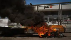 Colunas de fumaça preta se elevam de pneus em chamas em protesto do lado de fora dos portões fechados da FATE, em 18 de fevereiro de 2026.