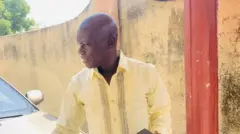 Di late Abdullahi Isah wear cream colour log sleeve shirt wey get buttons and broen design. E barb gorimakpa hair and dey look sideways as e keep hand ontop white car wey resemble Hilux