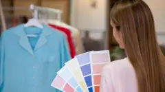 arkası dönük bir kadın elinde renk paletiyle askıdaki kıyafetlere bakıyor. 