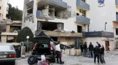 Várias pessoas estão do lado de fora de um prédio destruído, com as janelas estilhaçadas