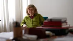 Svetlana Alexievich, sorridente, está sentada atrás de uma pilha de livros sobre sua mesa de trabalho, vestindo uma blusa verde