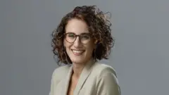 Ana Carolina Evangelista de cabelos cacheados curtos, óculos e terninho bege, sorrindo para a câmera