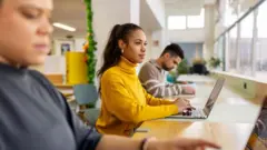 Stock photo shows students sitting at a table by a window in a student library working away at their computers.