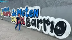 Em uma parede externa cinza de pé-direito duplo com arame farpado no topo, há uma placa com os dizeres “Se mexerem com Maduro, mexem com o bairro”, nas cores da bandeira venezuelana. Uma mulher caminha ao lado do mural.