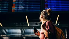 Mulher com celular olha para o quadro de partidas e chegadas de um aeroporto